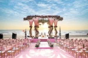 A romantic beach wedding setup at sunset with rows of pink and gold chairs facing a flower-decorated ceremony arch, a pink aisle runner, and floral arrangements overlooking the ocean waves under a partly cloudy sky.