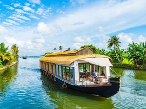 A traditional houseboat with a curved thatched roof cruises along a calm tropical backwater, surrounded by lush green palm trees and vegetation under a bright blue sky with scattered clouds.